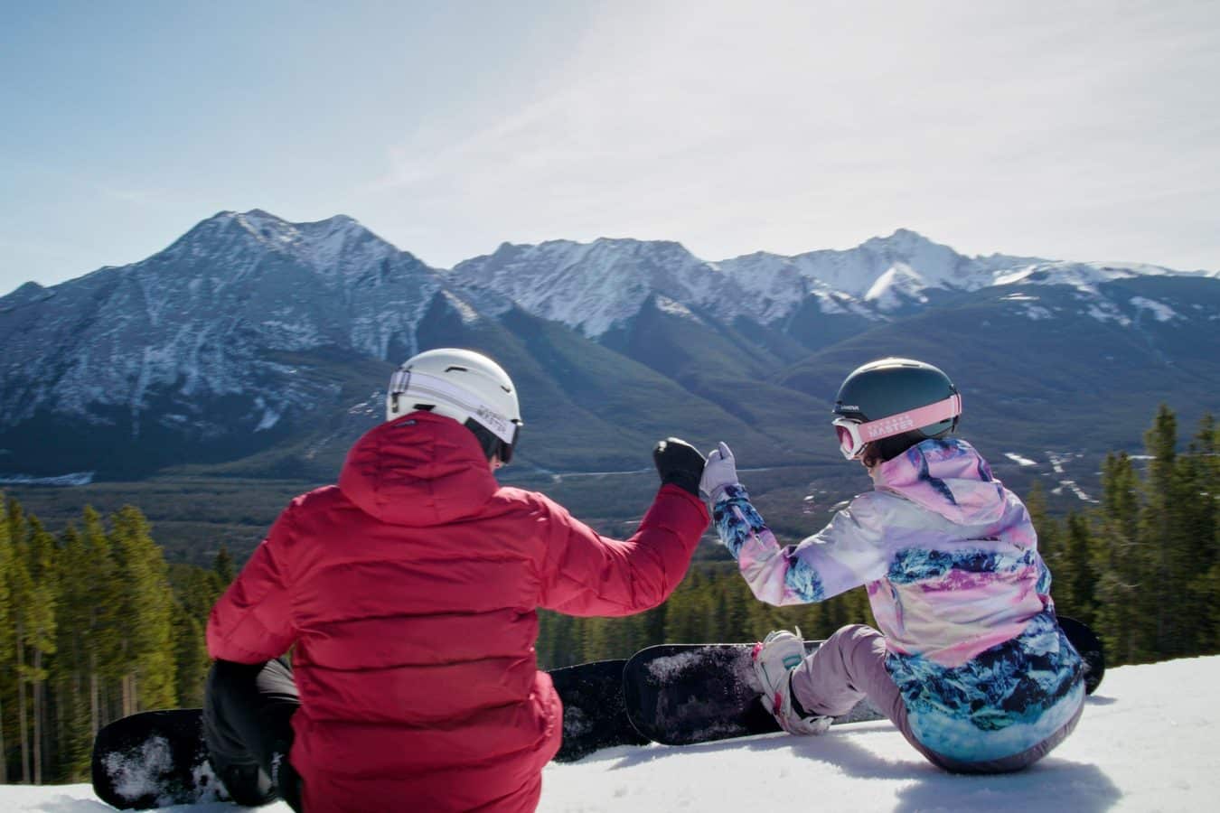 Two people in red and pastel ski jackets sitting on snow with snow covered mountains in the background'Tis the Season for Setups Review: Hallmark Kicks Off Countdown to Christmas 2026