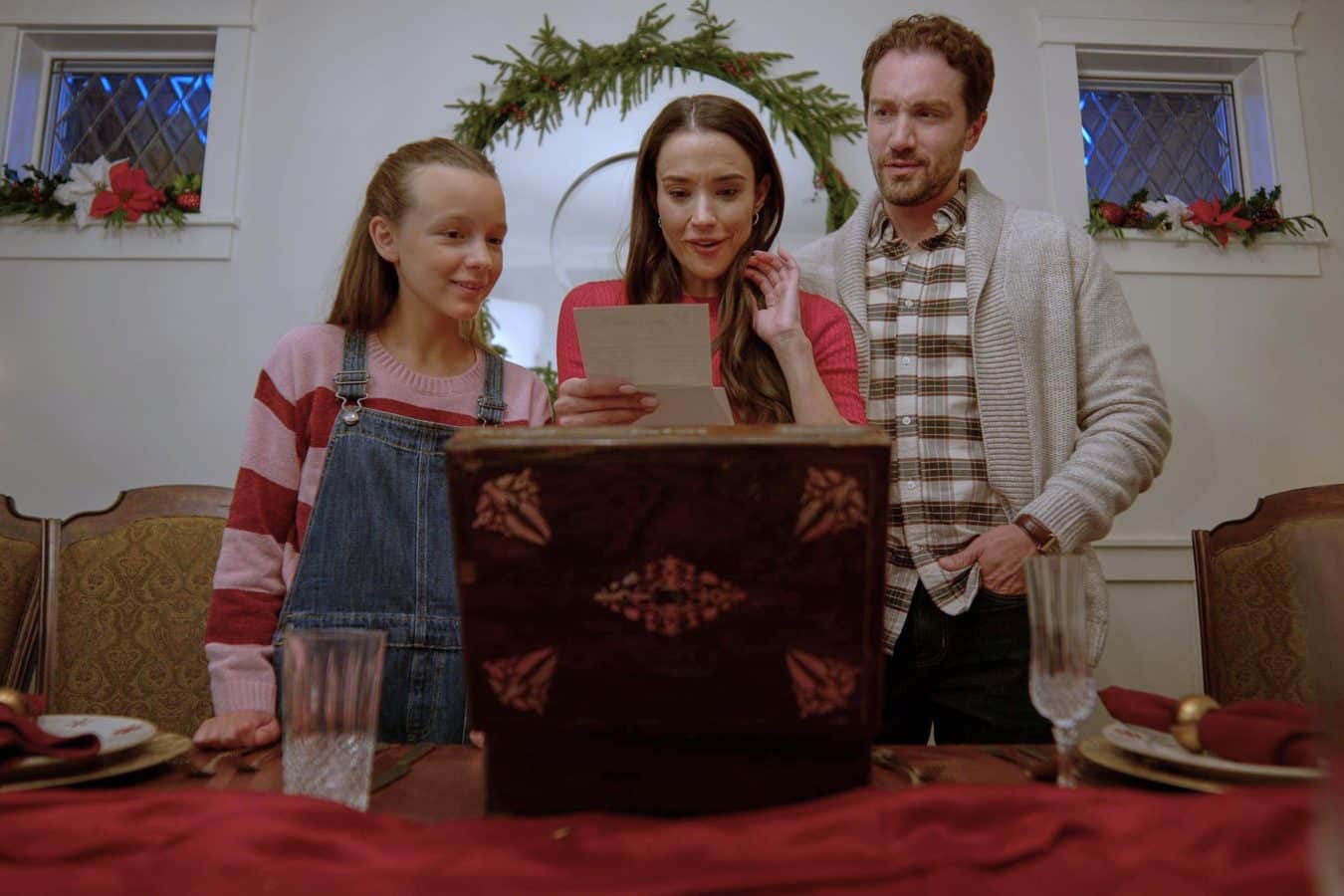 Family of three gathered around a table in a holiday setting reading a letter or card together as decorations loom in the background with garland and poinsettias