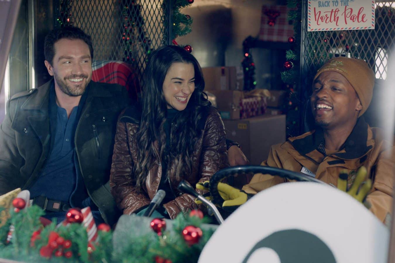 Three friends in a Christmas decorated vehicle sharing a laugh with garlands and a North Pole sign visible in the background