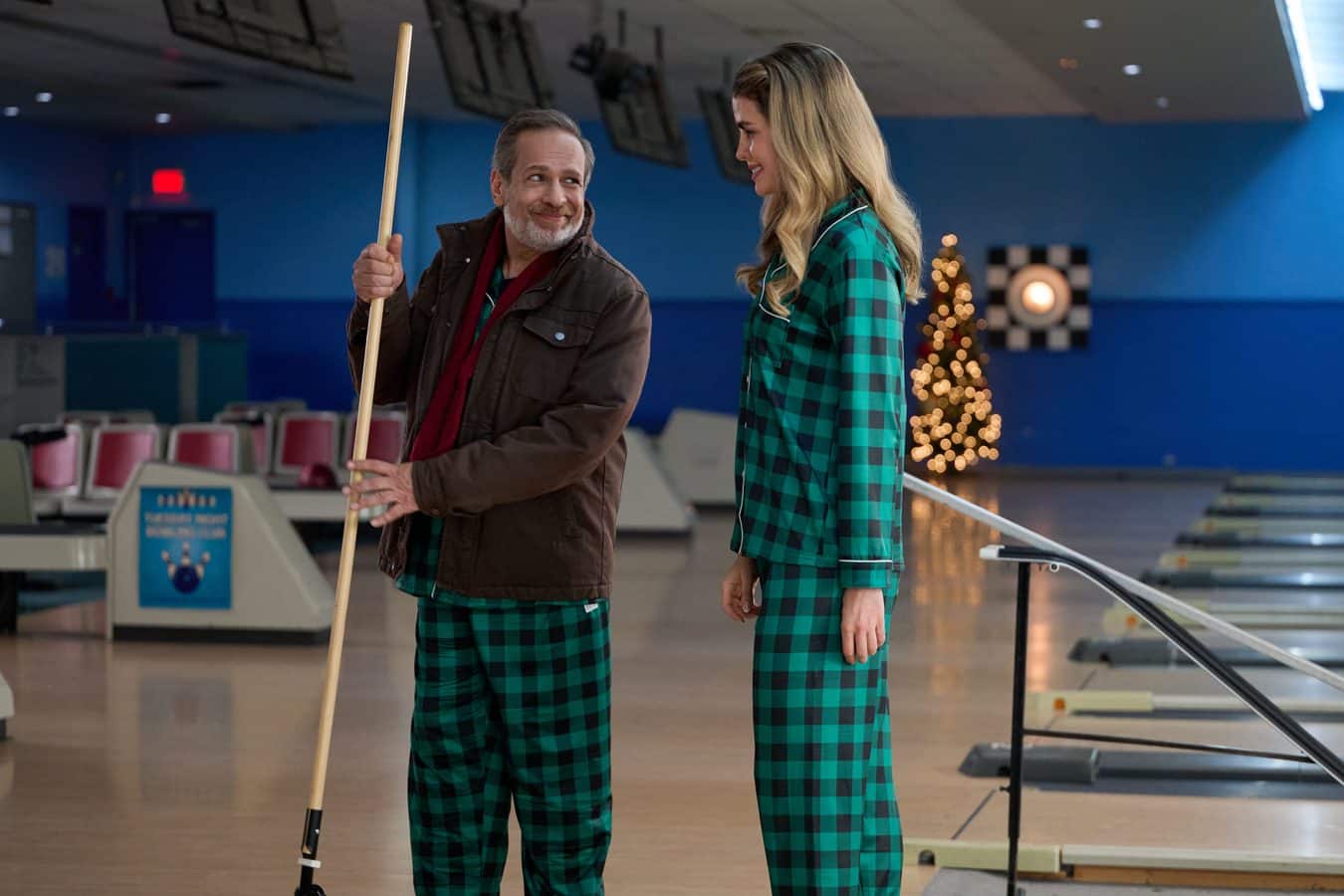 Ashley Newbrough as Audrey Bell shares a laugh with a co star in matching green plaid pajamas inside the bowling alley in Winter Wonderlanes